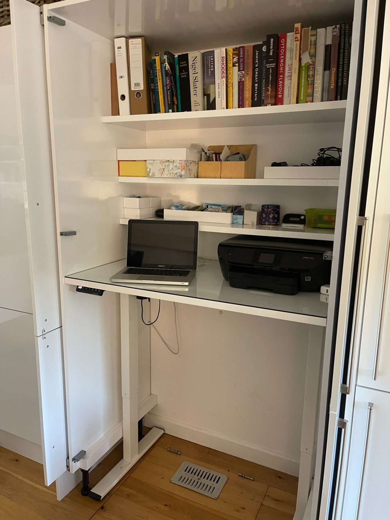 Desk riser set at standing position Compact workspace with a laptop and printer on a desk riser in a cupboard.