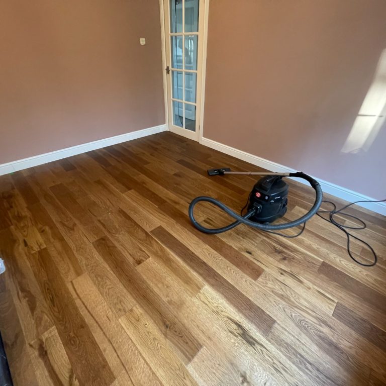 Picture of newly laid wooden floor with a vacuum cleaner in the corner and a glass door in the background.