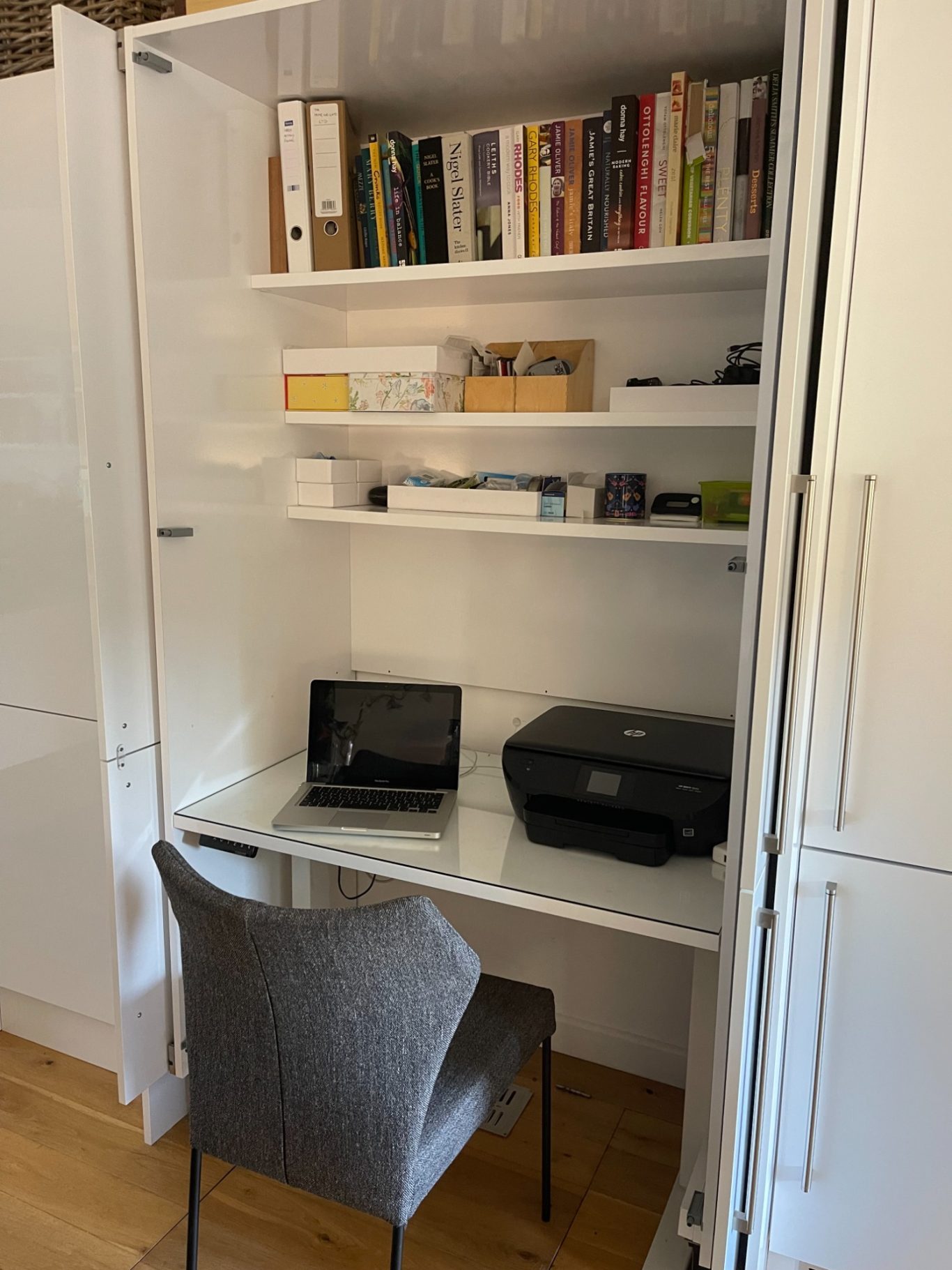 Desk riser set at seated hight Compact home office setup with laptop, printer, shelves, and a chair.